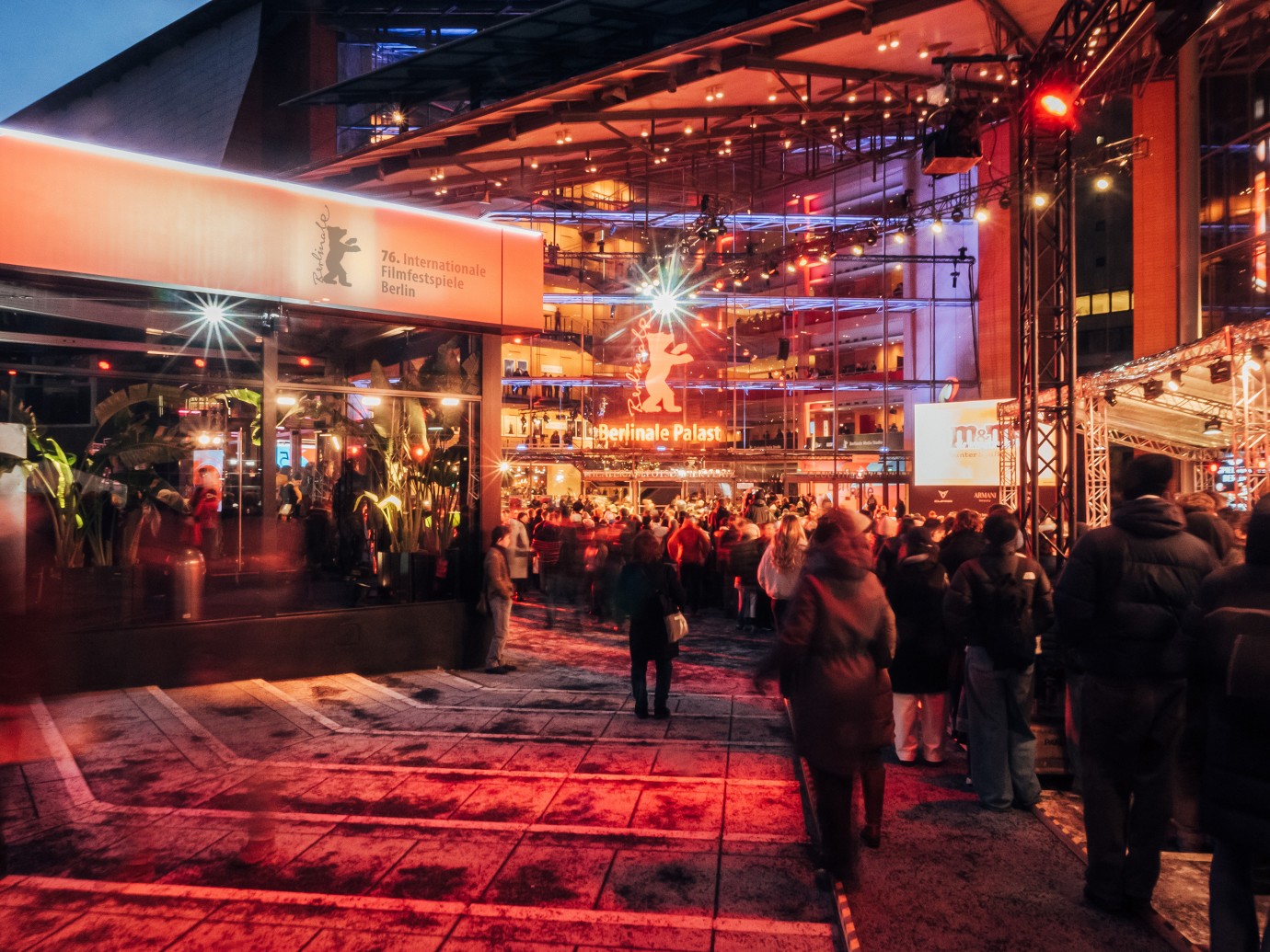 Crowd at a brightly lit Berlin film festival venue at night. Red and orange lights illuminate the bustling scene, creating a vibrant, festive atmosphere.