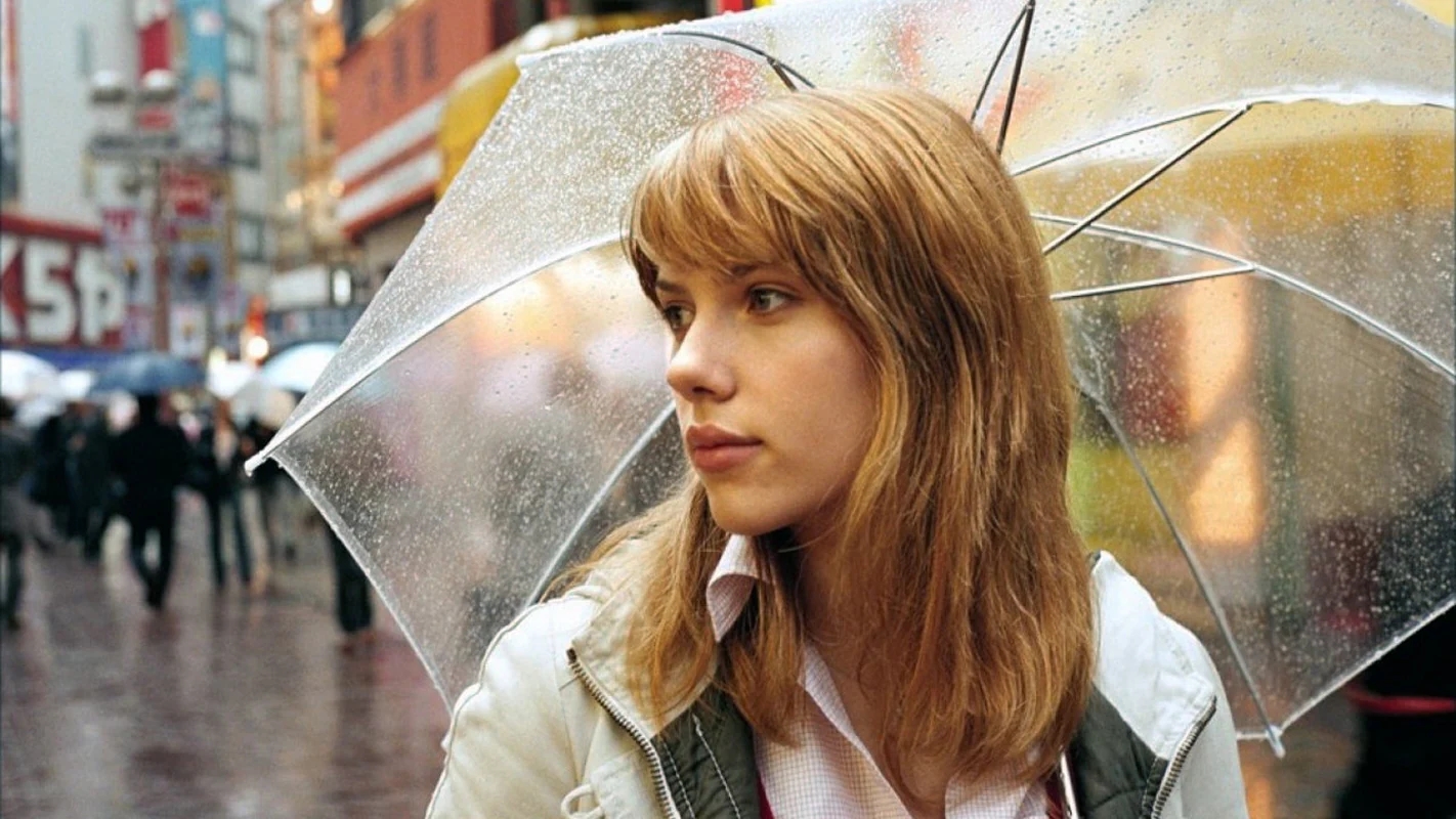 A person with shoulder-length blonde hair holds a clear umbrella in a bustling city street. The scene is rainy, with a soft, introspective mood.